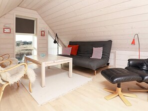 A cozy loft-style room with a wooden floor, a grey sofa with red and white cushions, a black leather chair, a wooden table, and a white rug. - 5 Star Holiday Home in Skagen (Skagen)