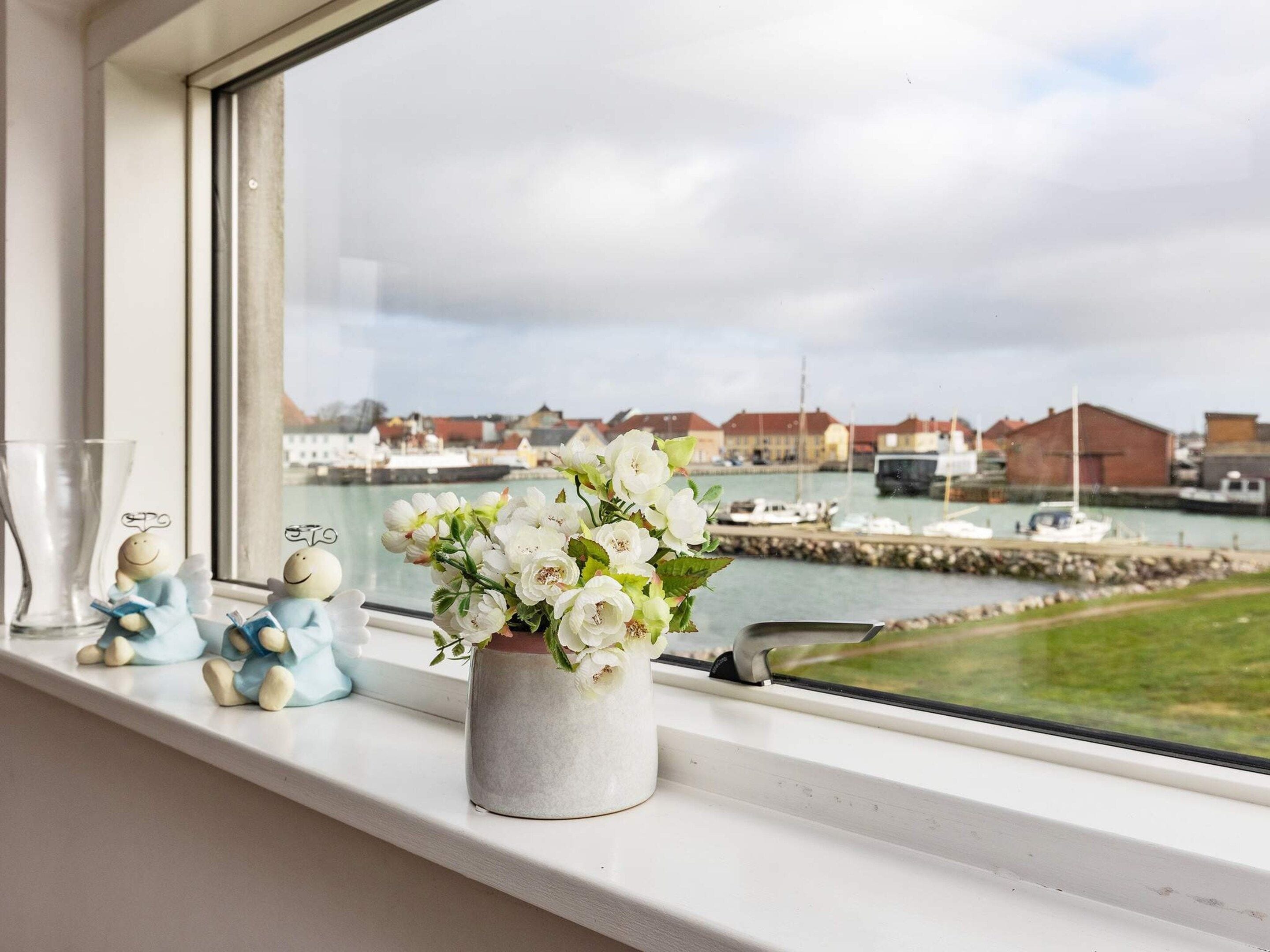 Una ventana con vista a un puerto deportivo, un florero con flores blancas y dos figuritas en el alféizar.