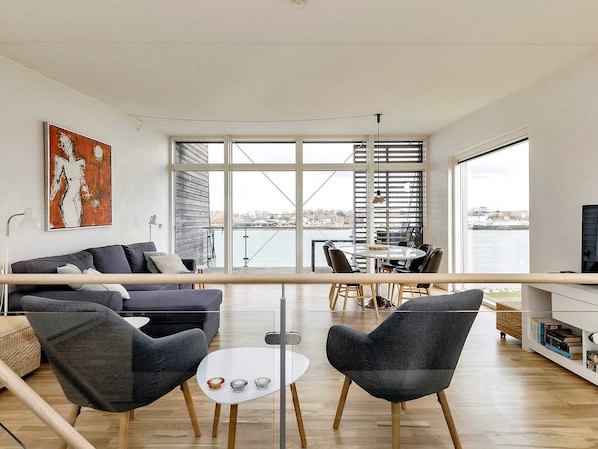 A modern living space with a sofa, chairs, a dining table, and a view of a body of water through large windows.