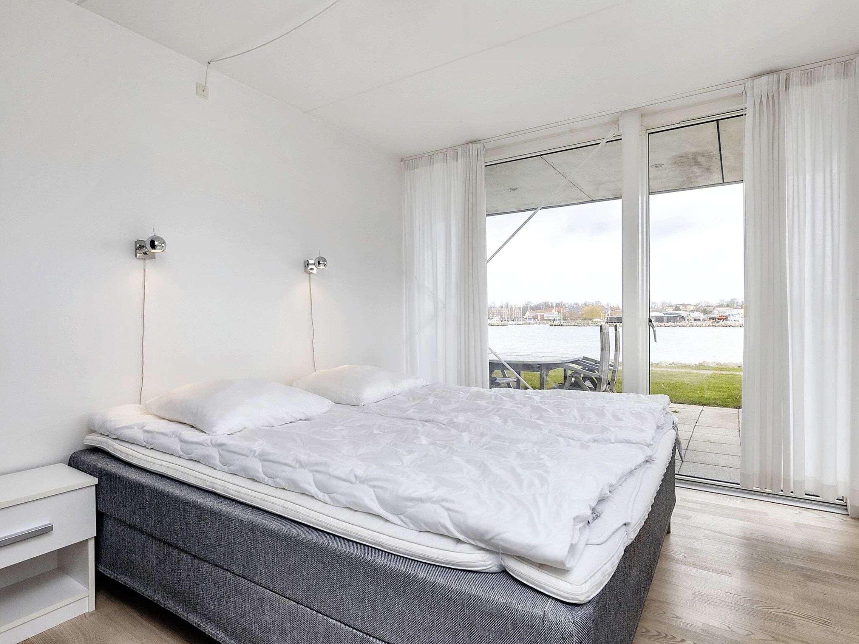 A bedroom with a bed, a nightstand, a wall-mounted light, and a view of a body of water through a sliding glass door.