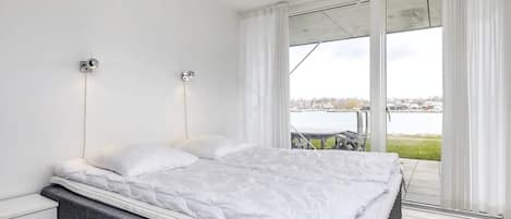 A bedroom with a bed, a nightstand, a wall-mounted light, and a view of a body of water through a sliding glass door.