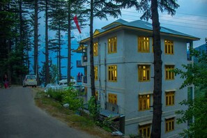 Un edificio de varios pisos con estilo arquitectónico tradicional, rodeado de altos árboles y un paisaje montañoso.