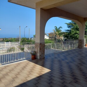 Ein überdachter Außenbereich mit gefliestem Boden, einem weißen Metallzaun und Blick auf das Meer.