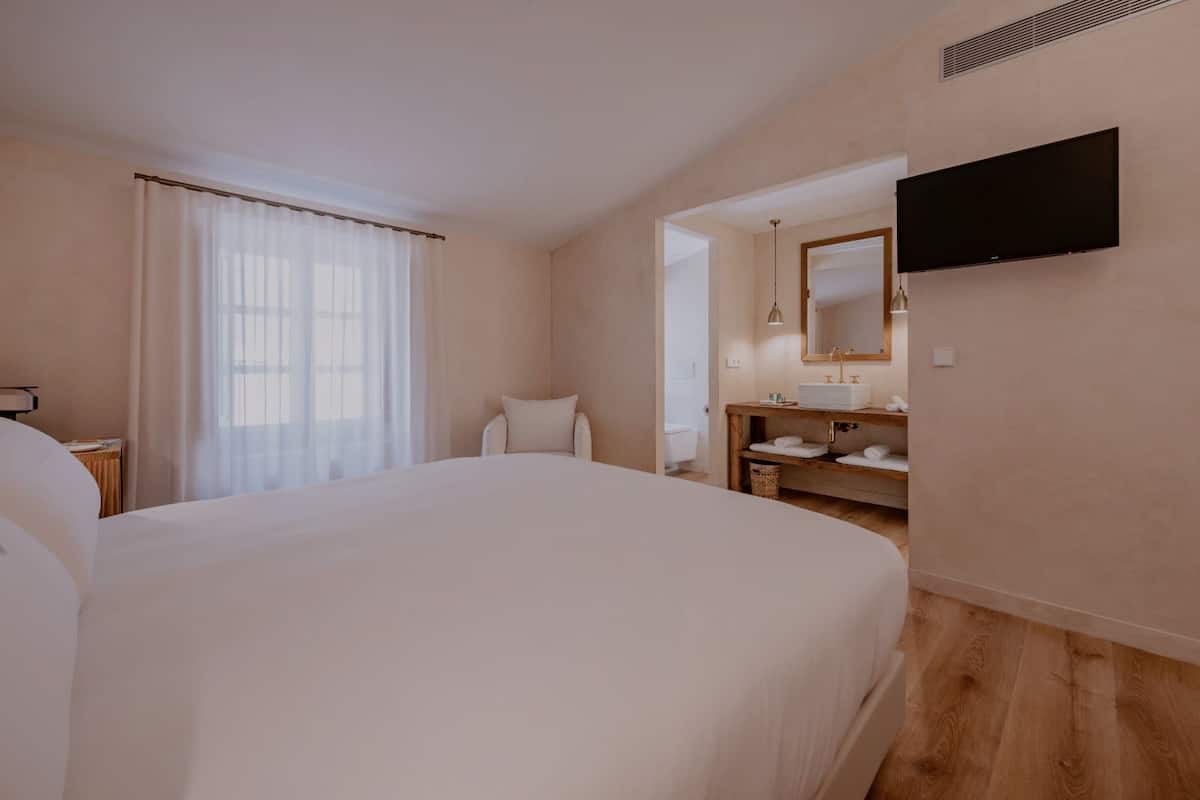 Habitación de hotel con una cama grande, suelo de madera, baño con lavamanos y espejo, y un televisor montado en la pared.