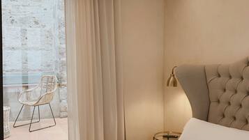 Ein modernes Hotelzimmer mit einem großen Bett, einem kleinen Nachttisch, einer Lampe und Blick auf einen Balkon mit einem Korbstuhl.