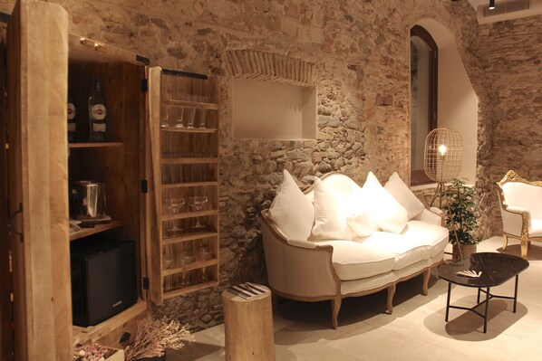 A cozy living room with a stone wall, a white sofa, a wooden cabinet with glassware, and a small table with a plant.