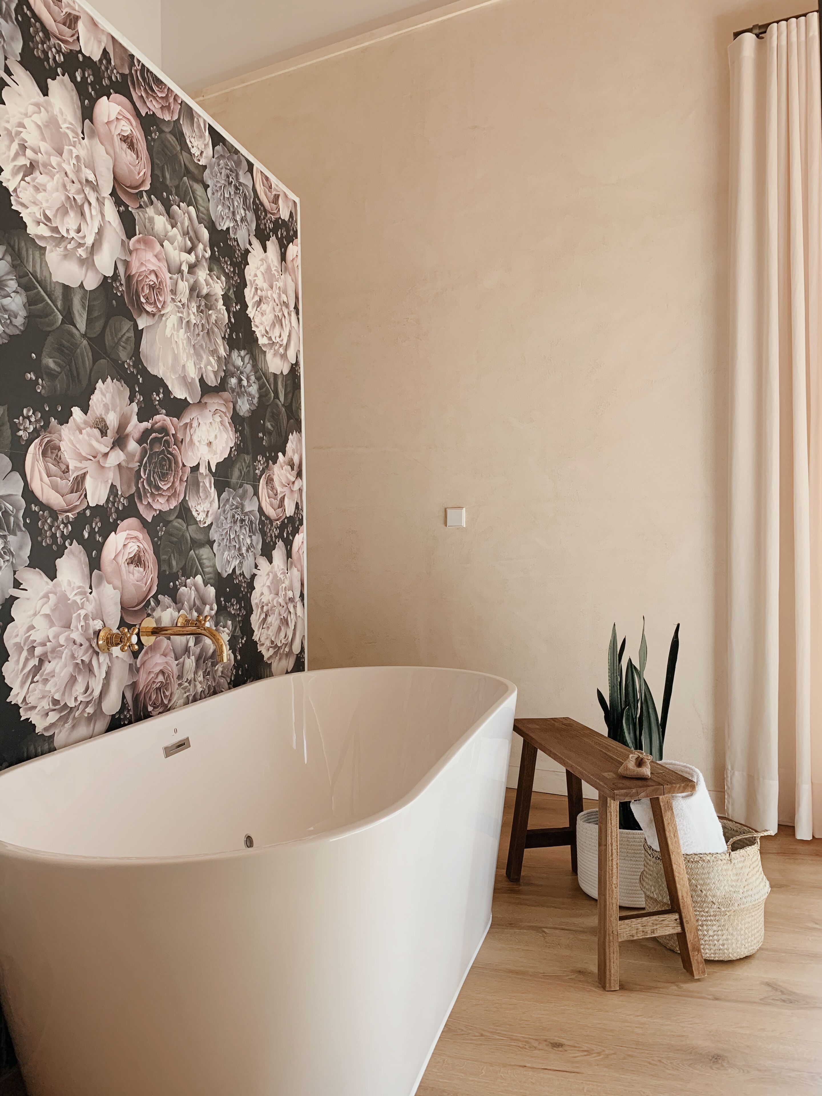Ein Badezimmer mit einer freistehenden Badewanne, einer Holzbank, einer Topfpflanze und einem floralen Wanddesign.