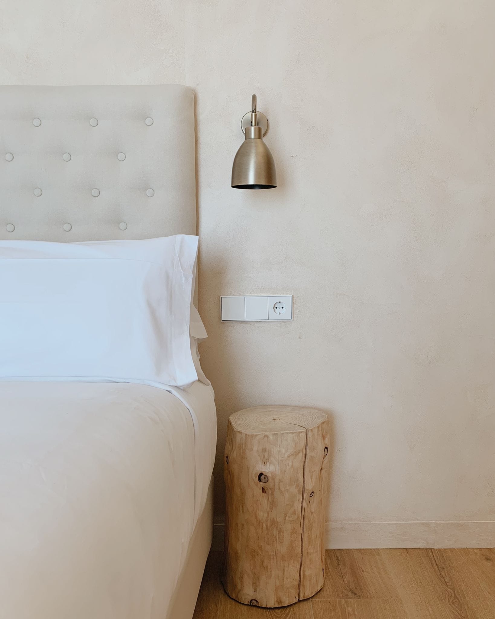 Un dormitorio con una cama, una lámpara de noche, un enchufe y un taburete de madera.