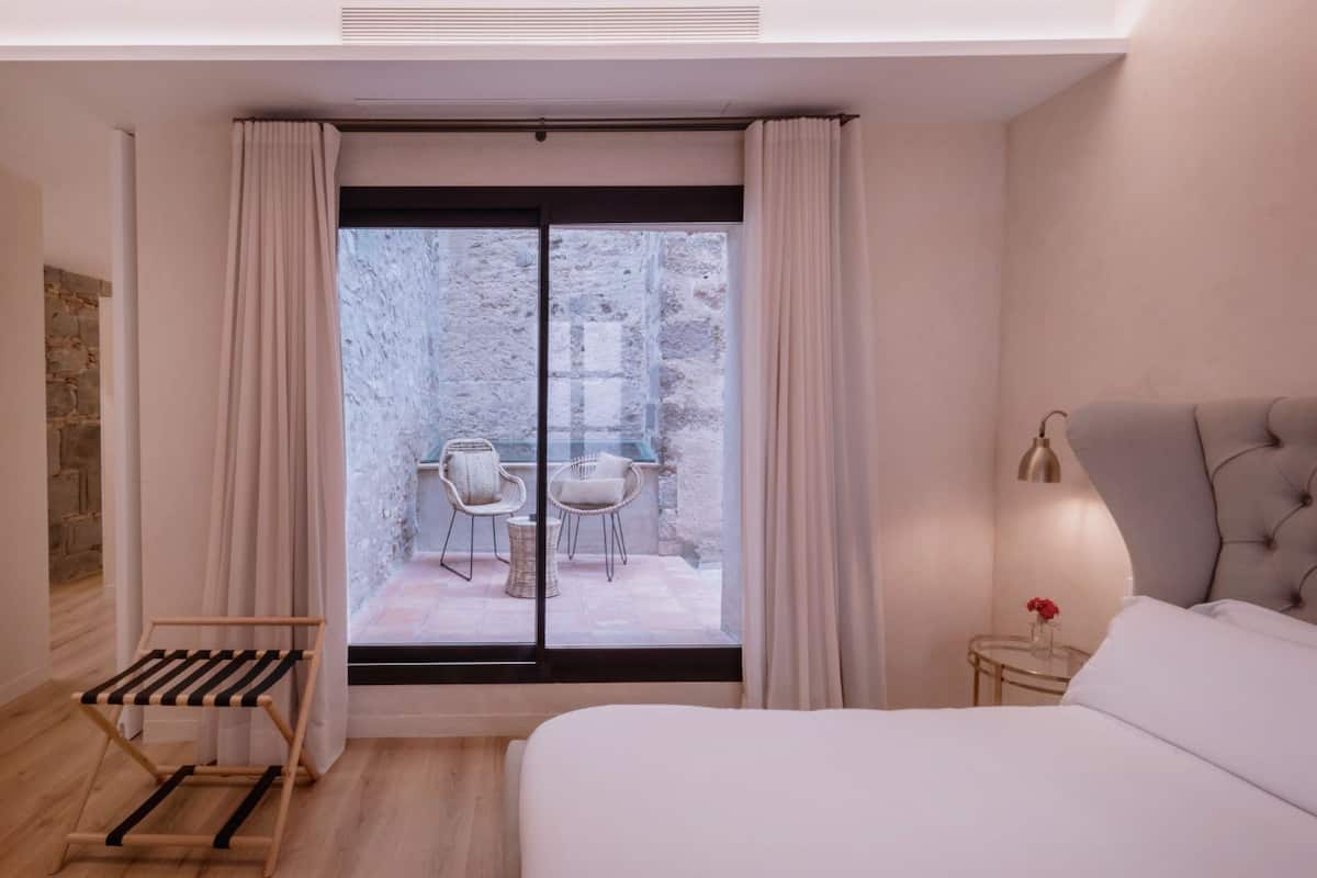 Un dormitorio moderno con una cama grande, una mesita de noche con lámpara, una silla y vistas a un patio exterior con mesa y sillas.