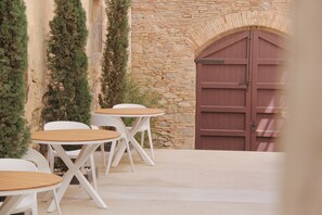 Un patio con mesas redondas y sillas blancas, un muro de piedra y una puerta de madera.