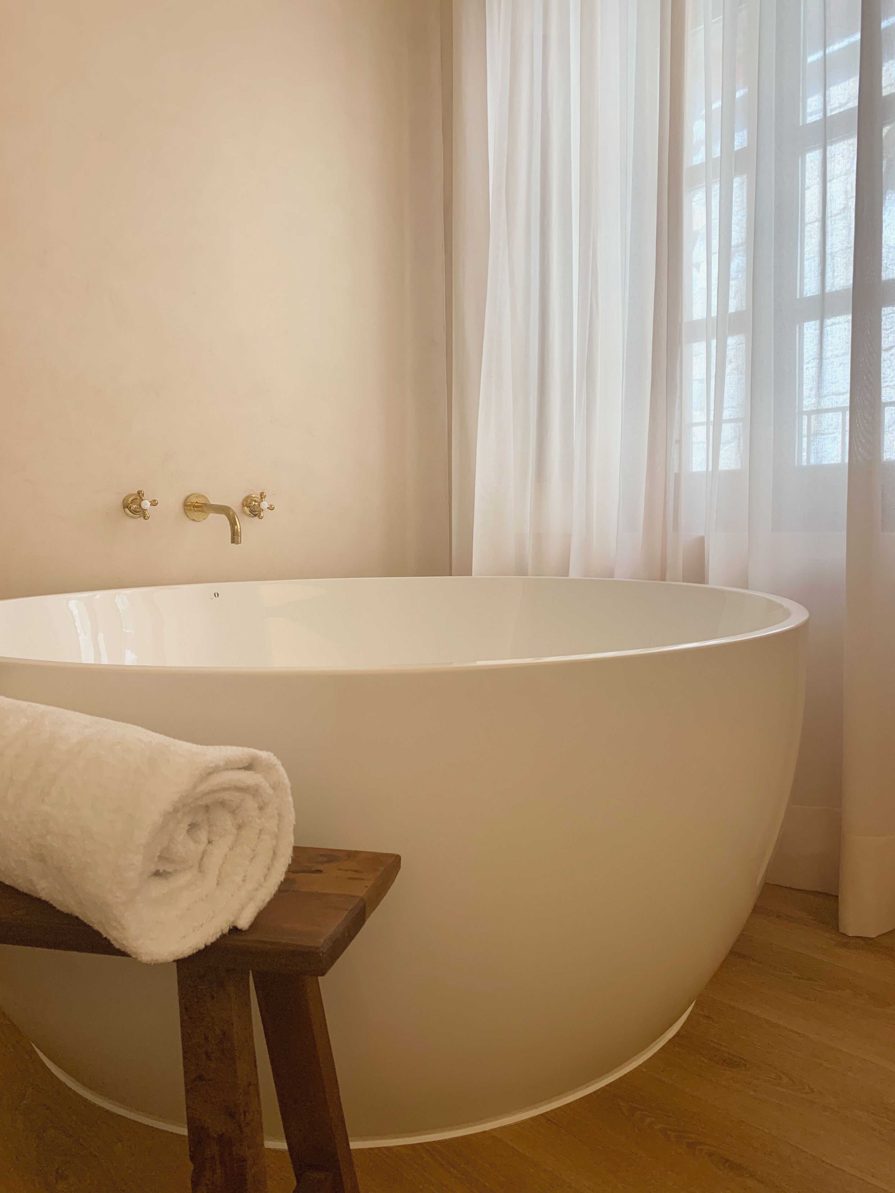 A large freestanding bathtub in a bathroom with a wooden stool and a rolled towel beside it.