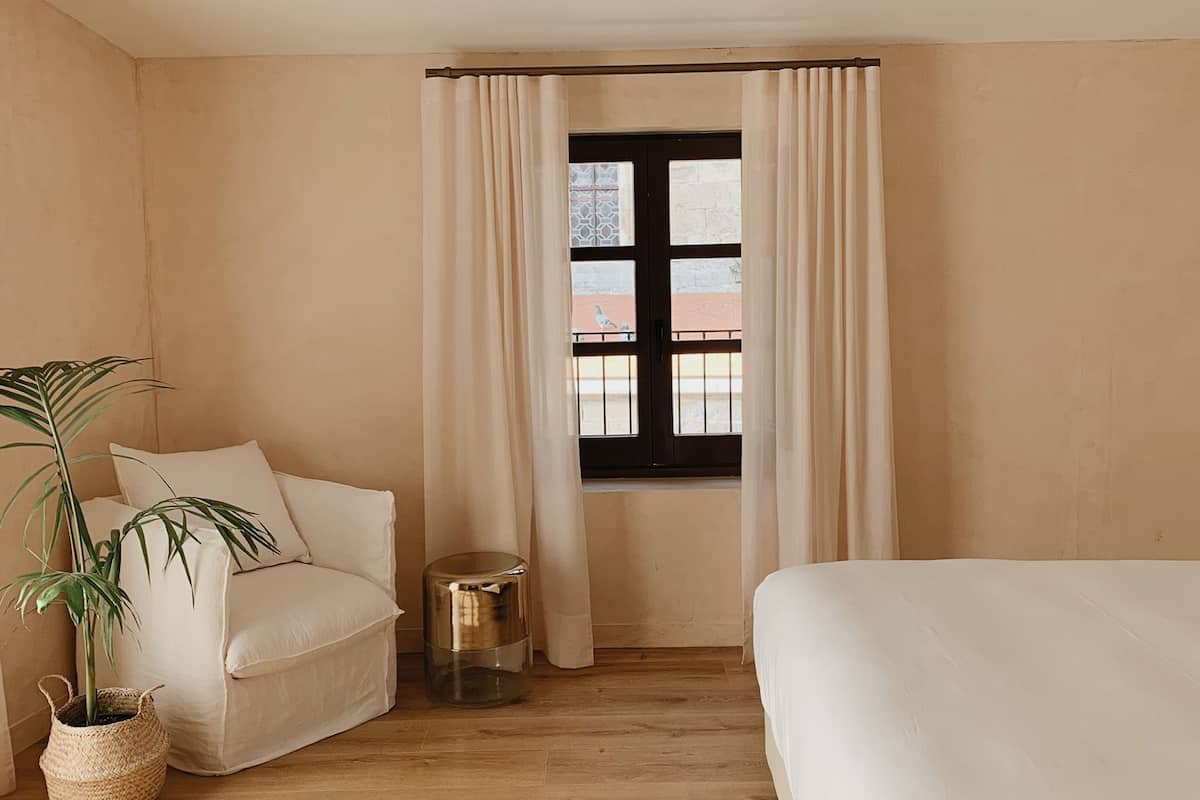 Un dormitorio minimalista con suelo de madera, una cama, un sofá, una ventana con cortinas y una planta en maceta.