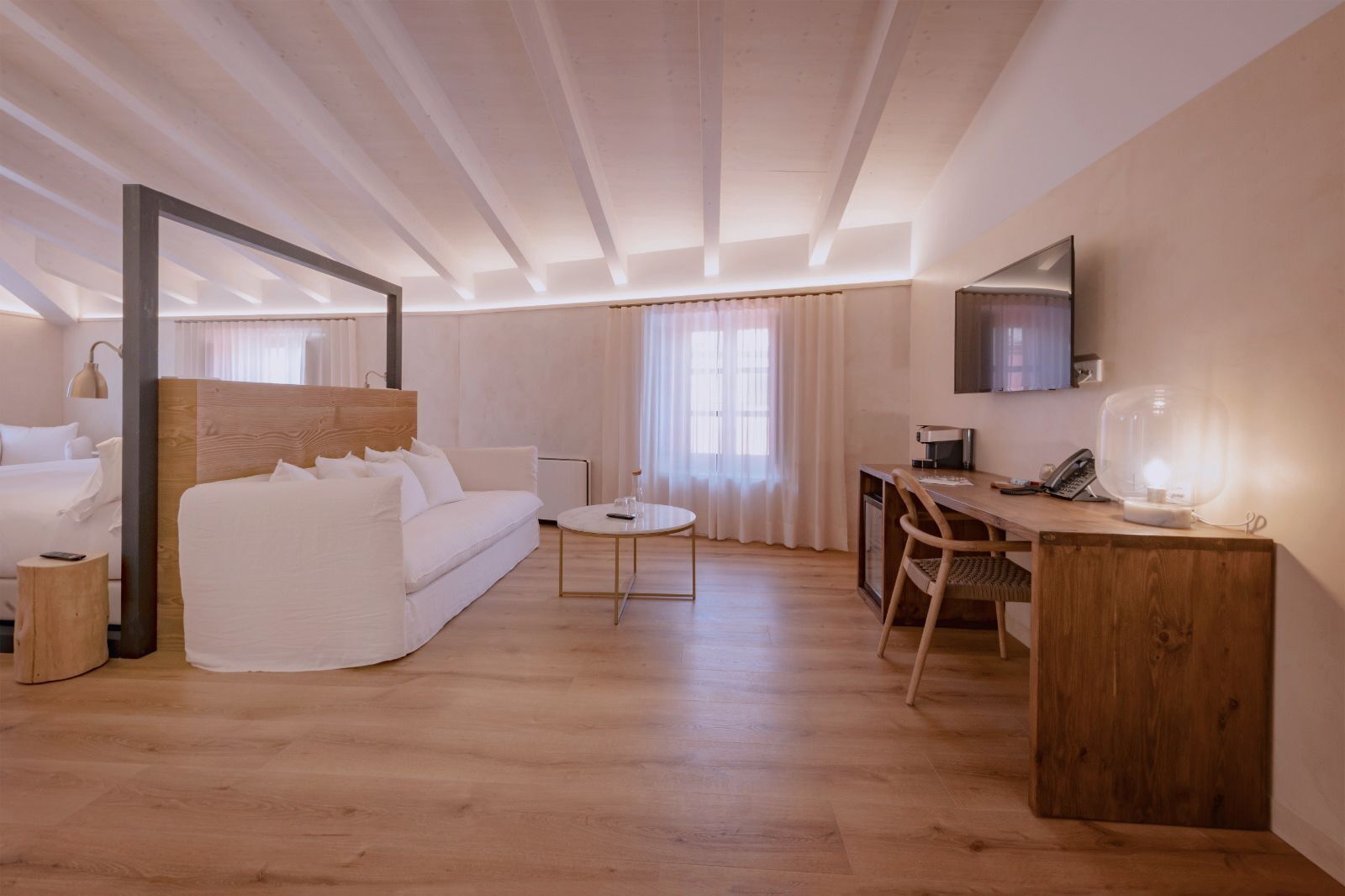 Habitación de hotel moderna con techo de madera, sofá blanco, una pequeña mesa redonda, escritorio con silla, espejo y televisor montado en la pared.