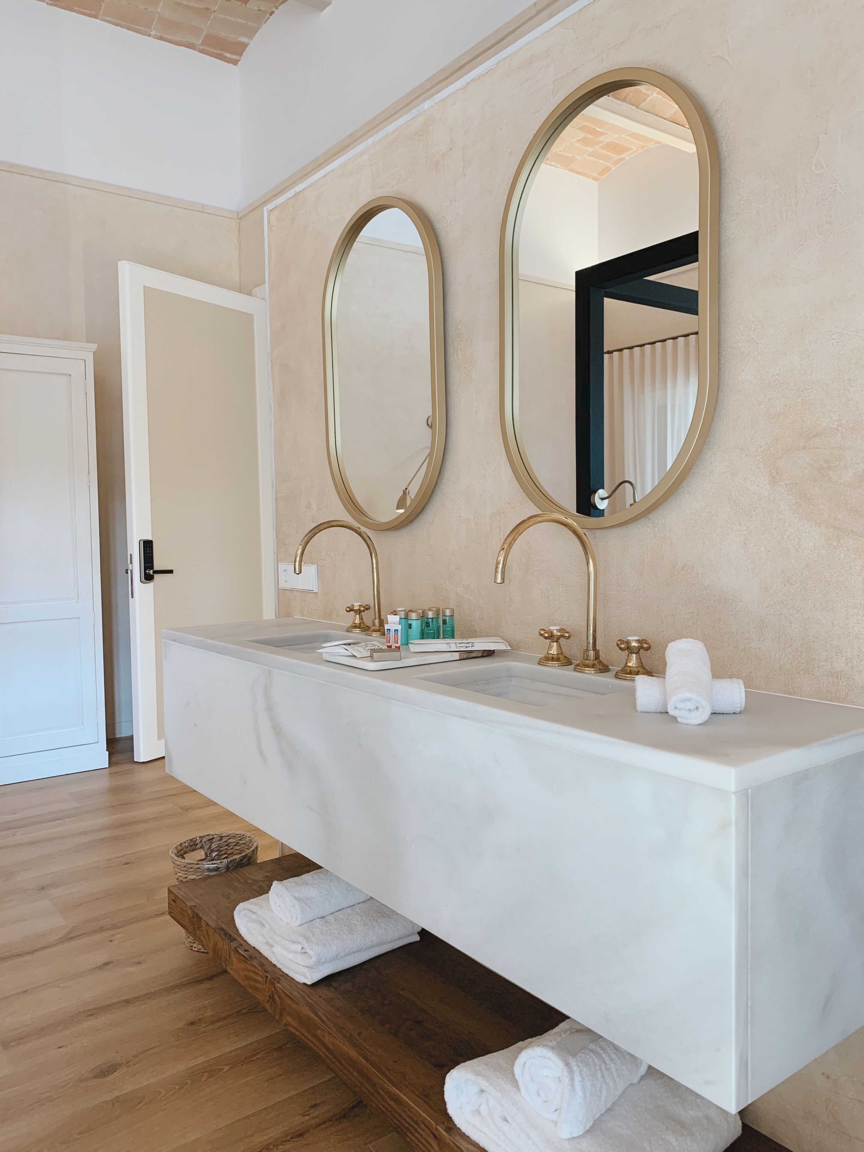 Un baño moderno con un tocador amplio, dos espejos con marcos dorados, un lavamanos blanco y un estante de madera con toallas.