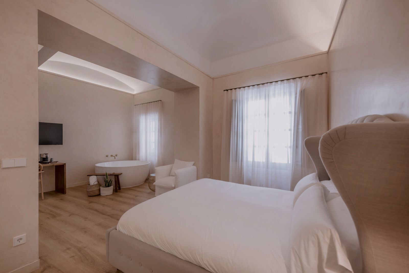 A modern hotel room with a large bed, a desk, a chair, a bathtub, and a TV mounted on the wall.