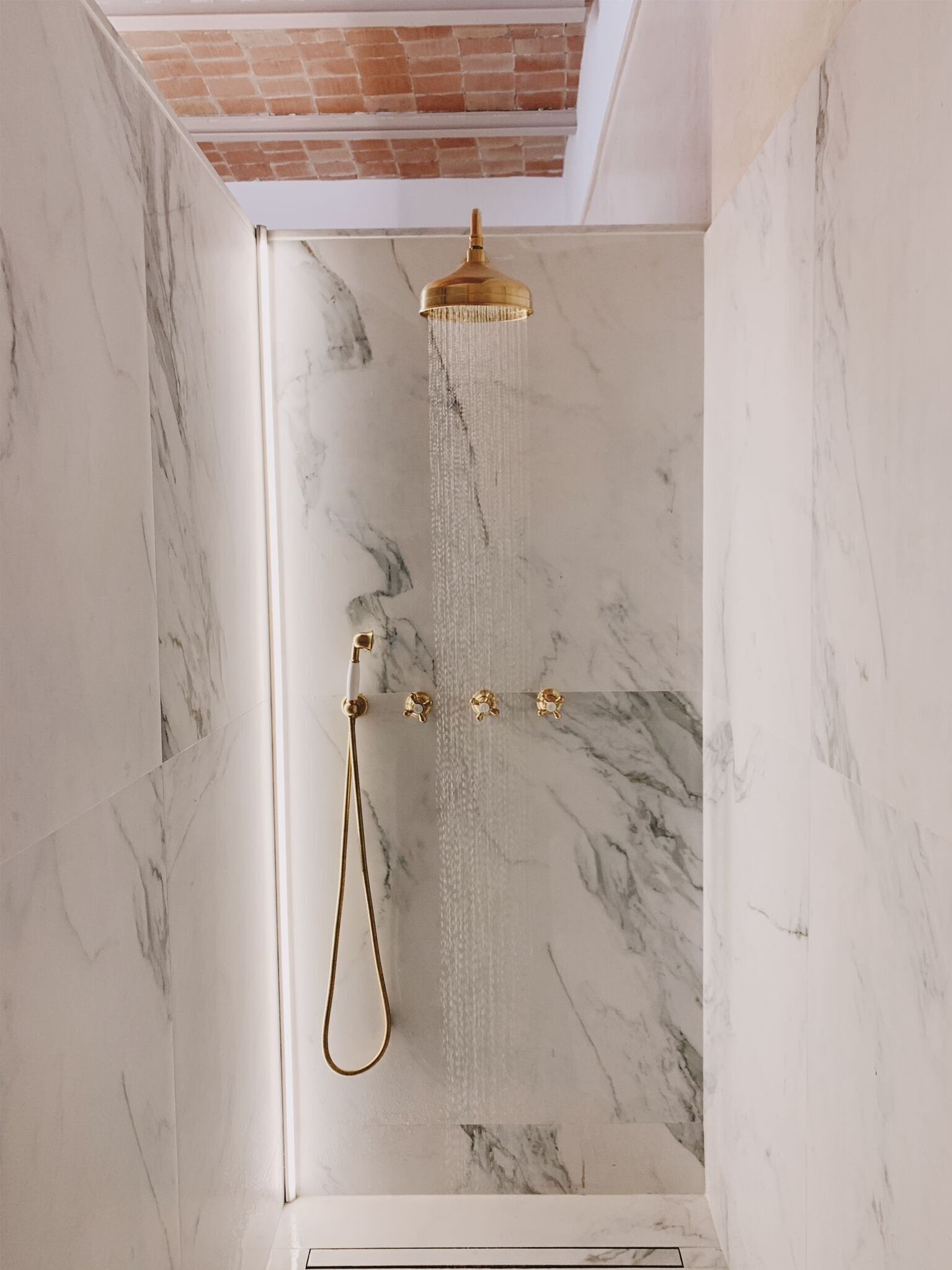 Ducha con cabezal dorado y ducha de mano frente a una pared de mármol.