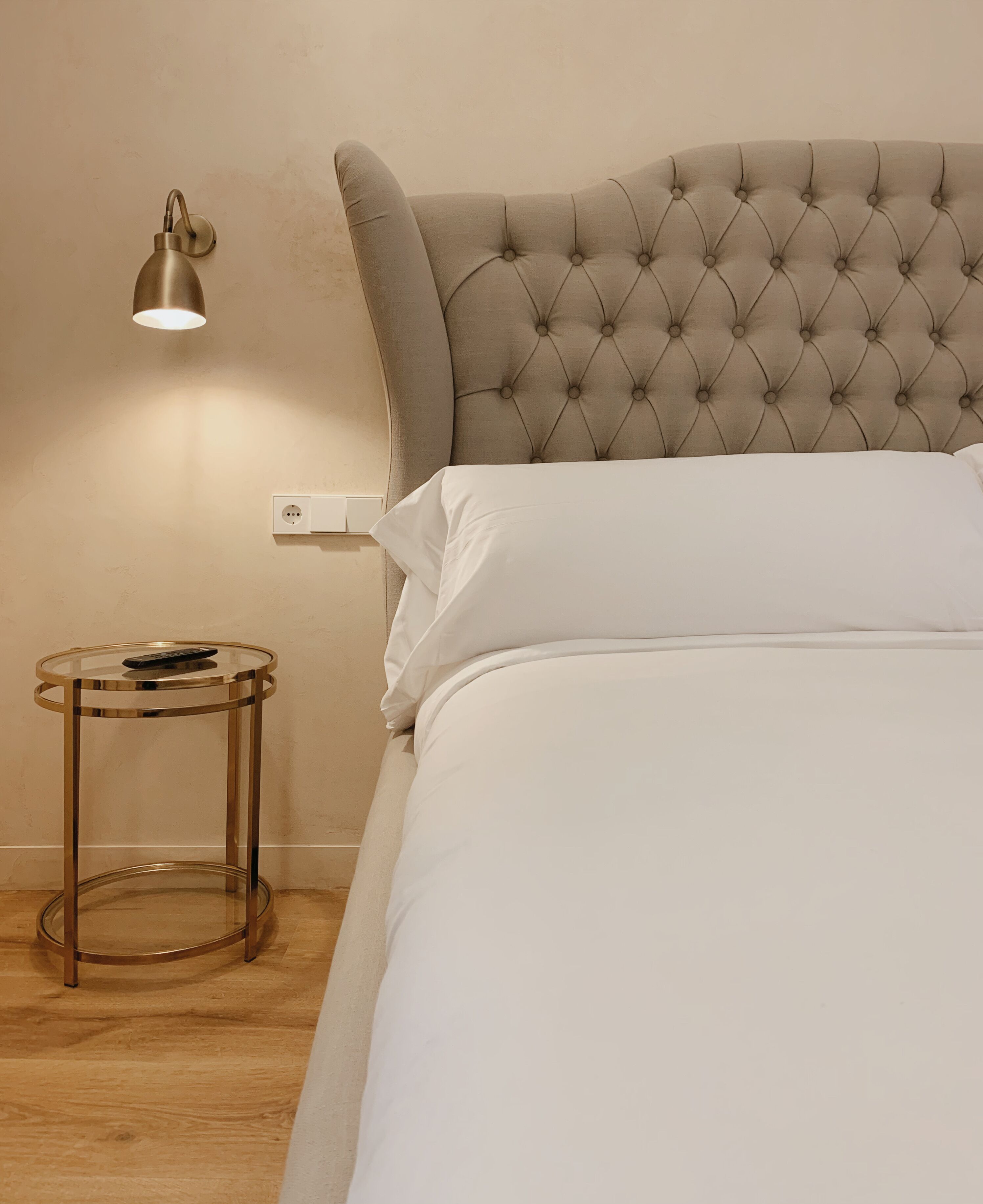 A neatly made bed with a tufted headboard, a bedside table with a lamp, and a power outlet on the wall.