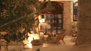 A cozy living room with a sofa, chairs, and a lamp. There is a large window with a view of the outside. - Palau Macelli (Castello d'Empuries)