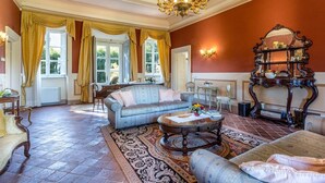 A spacious living room with a large sofa, a coffee table, and a chandelier. There are two windows with curtains, a fireplace, and a wooden sideboard with a mirror.