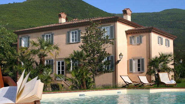 A large building with a swimming pool, surrounded by palm trees and a mountain in the background.