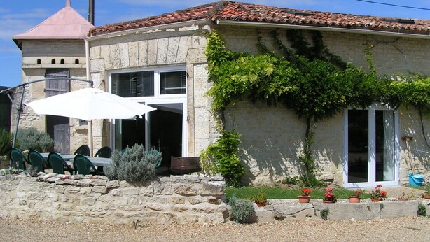 Un edificio de piedra con tejado de baldosas, una pequeña torre y un patio con sillas y una sombrilla.