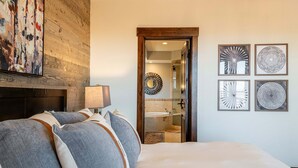 Quarto com cama, almofadas, abajur, lareira e banheiro visível através de uma porta de vidro.