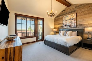 A bedroom with a large bed, a wooden headboard, a chandelier, a painting, and a view through a large window.