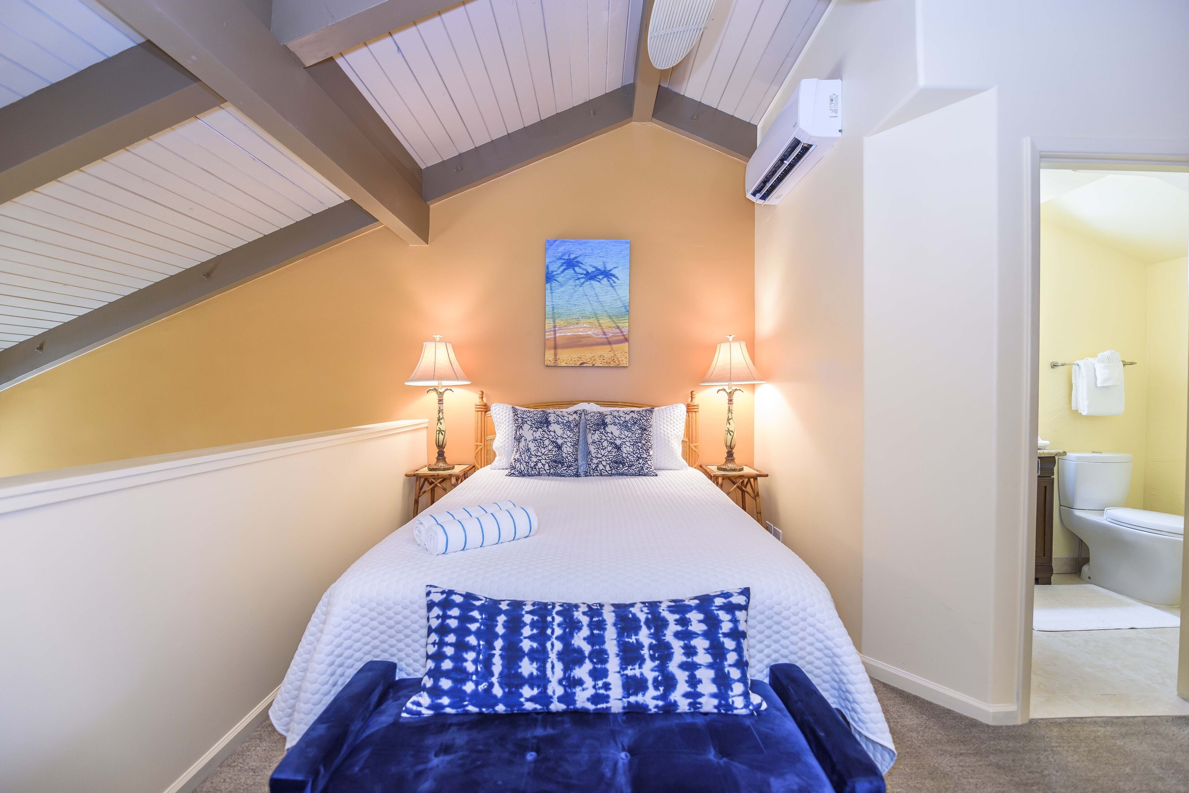 Quarto com uma cama grande, duas luminárias de cabeceira, um quadro na parede e vista para o banheiro.