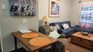 A living room with a wooden dining table, a sofa, and a coffee table. There is a painting of pelicans on the wall and a framed picture above the sofa. - Dauphin Island Beach Club 310B Budget Friendly near the Beach!! (Dauphin Island)