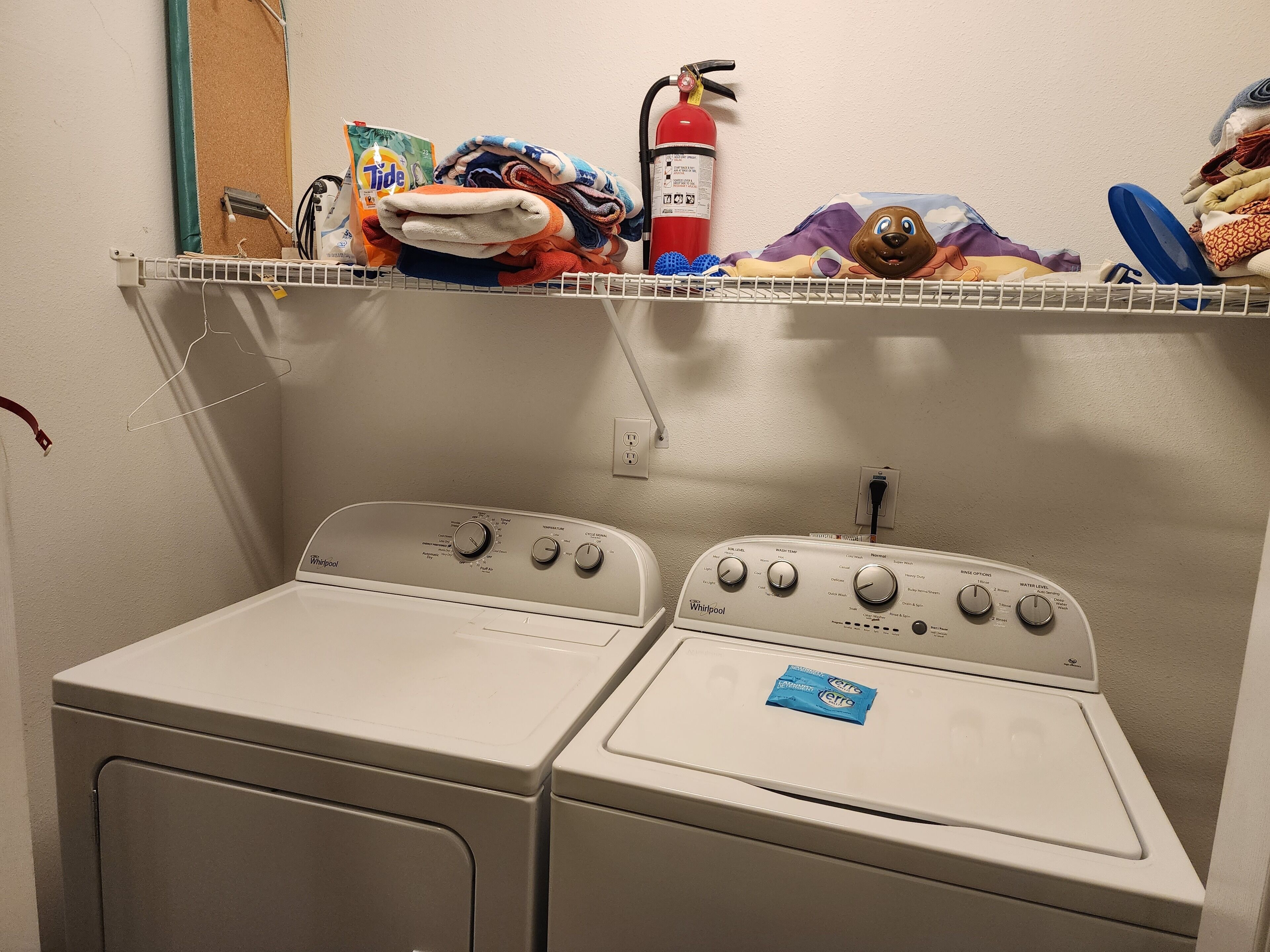 Een wasruimte met twee wasmachines en een plank erboven met opgevouwen kleding, een brandblusser en een zak Tide wasmiddel.