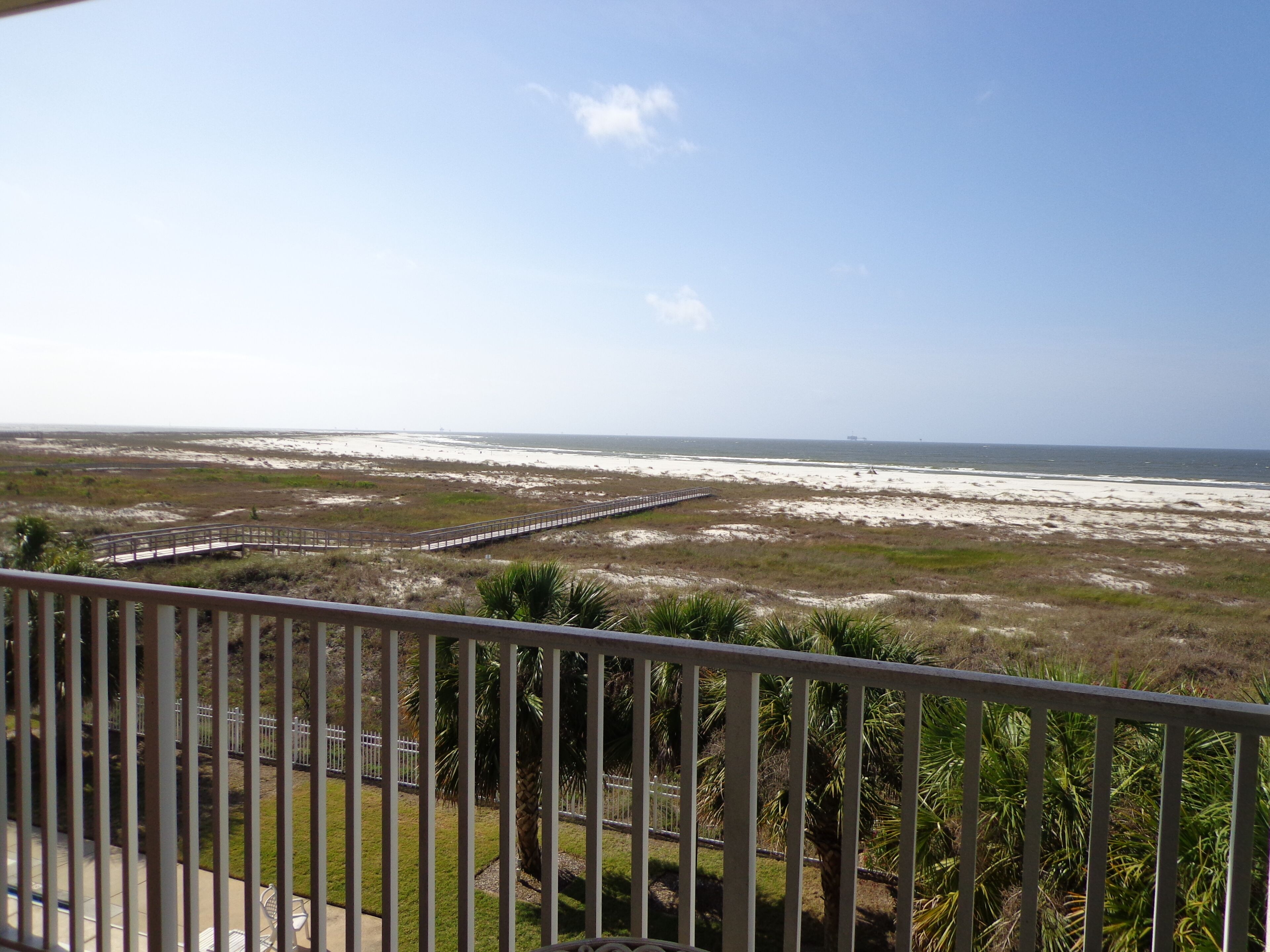 A view of a beach with a wooden boardwalk leading to the shore, surrounded by grassy dunes and palm trees.
