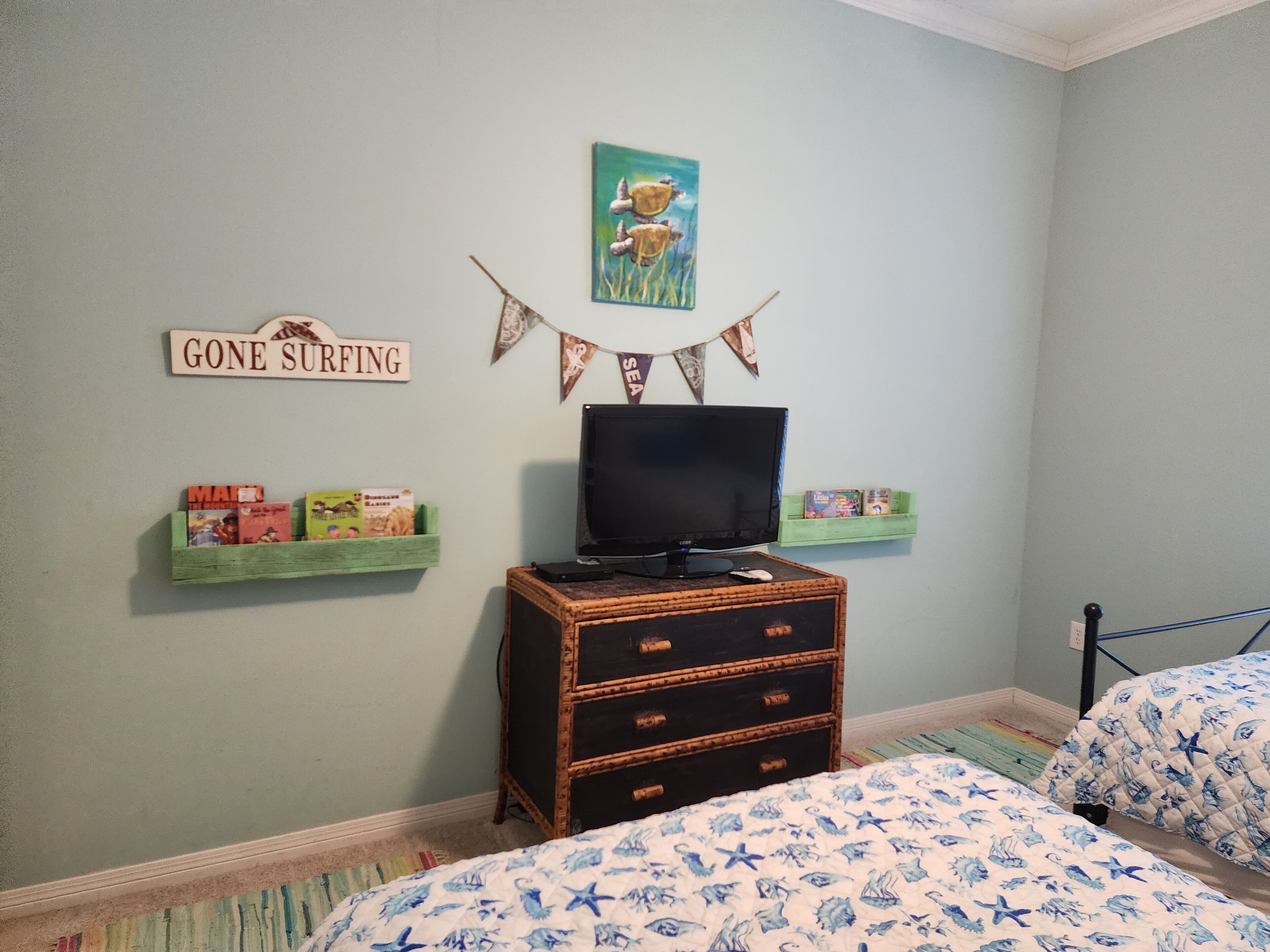 A bedroom with two beds, a dresser, a television, and wall decorations including a 'Gone Surfing' sign and a picture of a bird.