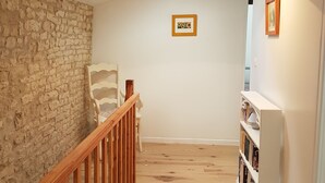 Un pasillo estrecho con piso de madera, una pared de piedra a un lado y una pared blanca al otro. A la izquierda, una barandilla de madera, una silla blanca y una estantería con libros y una lámpara.
