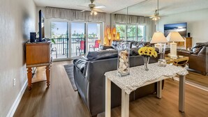 A modern living room with a leather sofa, a coffee table, and a TV. There is a balcony with a view of palm trees and a cityscape. - Land's End 305 building 3 ~ Beautifully remodeled ~ Across from Pool (Treasure Island)
