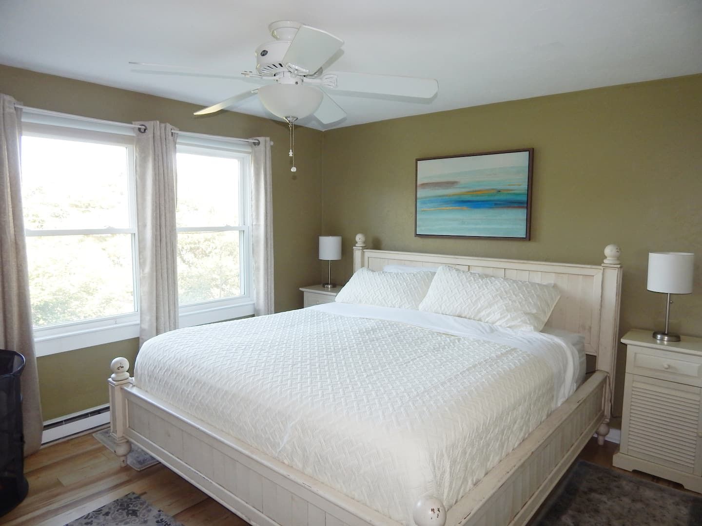 Une chambre à coucher avec un lit, un ventilateur de plafond, une fenêtre avec des rideaux, une table de chevet avec une lampe et un tableau au mur.