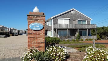 Une maison à deux étages avec un balcon au deuxième étage, un pilier en briques portant un panneau de signalisation et une allée gravillonnée.