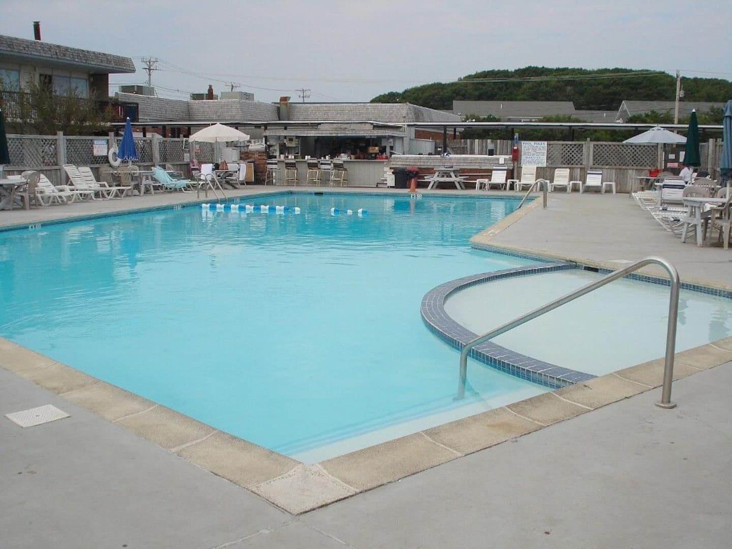 Piscina com trampolim, cadeiras de descanso e um pequeno edifício ao fundo.