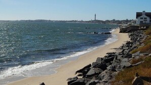 Ein Küstenstrand mit felsigem Ufer, ein weißes Gebäude mit dunklem Dach und ein Leuchtturm in der Ferne.