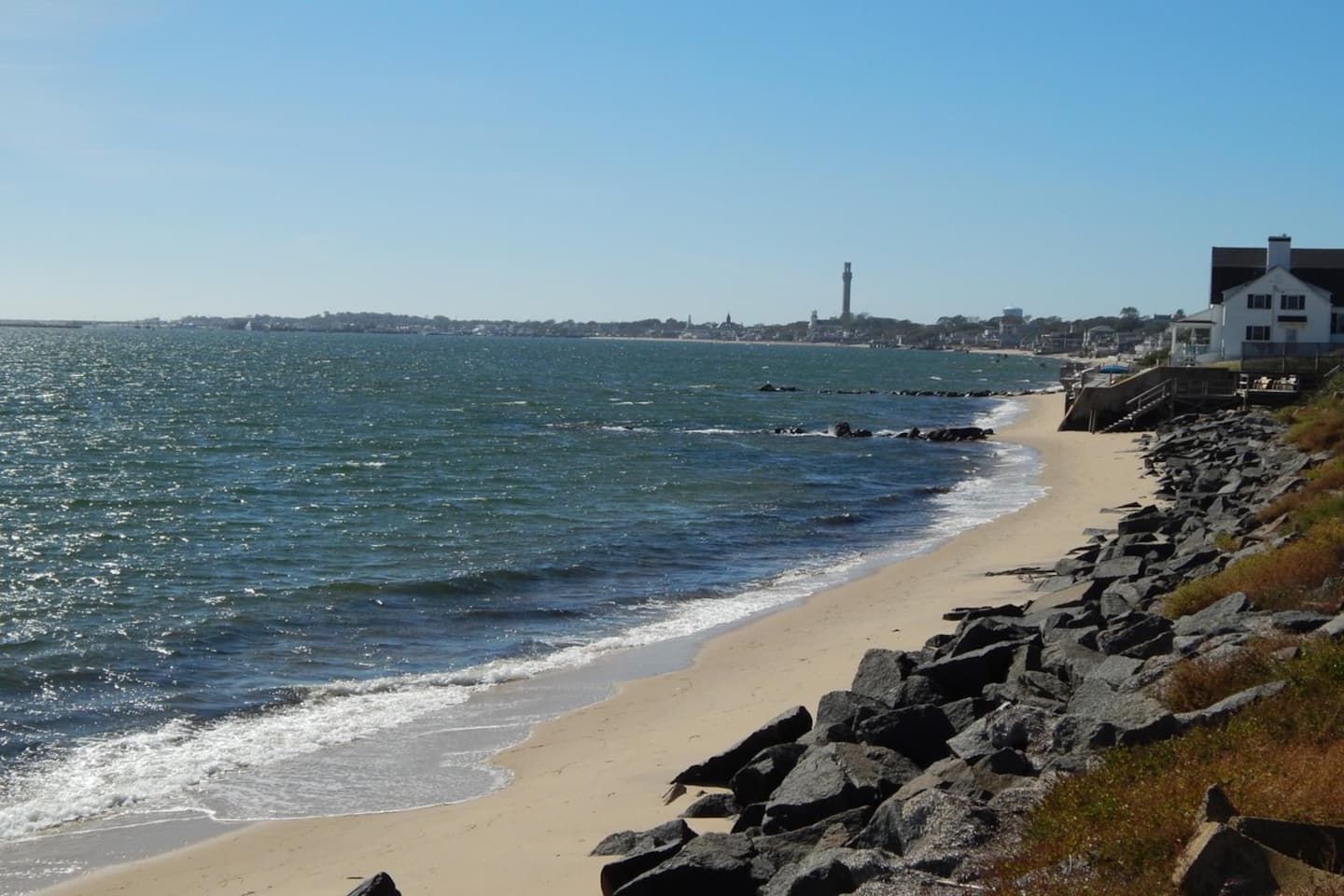 A coastal beach with a rocky shoreline, a clear sky, and a distant cityscape.