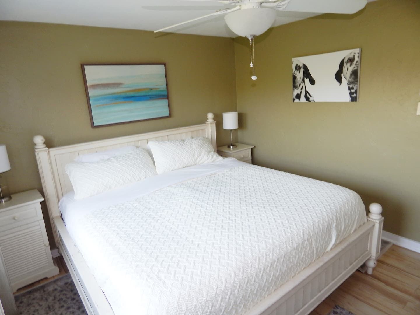 Quarto com cama, mesa de cabeceira, luminária, ventoinha no teto e duas obras de arte nas paredes.
