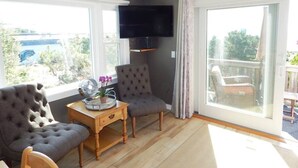 Ein Wohnzimmer mit grauem Sofa, einem kleinen Tisch mit Ventilator und Blumen, einem Fernseher an der Wand und einem Balkon mit Blick auf Grünflächen und eine blaue Struktur.