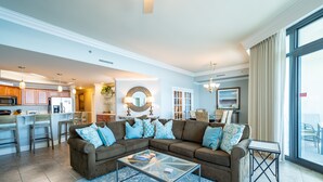 A modern living room with a sectional sofa, a glass coffee table, and a view of the kitchen and dining area.