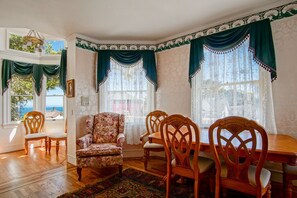 A dining room with a wooden table and chairs, a floral armchair, and large windows with green curtains. - Daffodil House - Pacific Grove House - Block to the Beach (Pacific Grove)