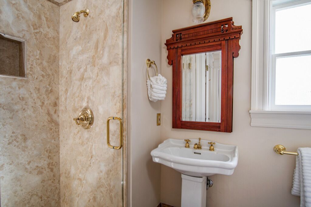 A bathroom with a marble shower, a white sink with gold fixtures, and a wooden-framed mirror.