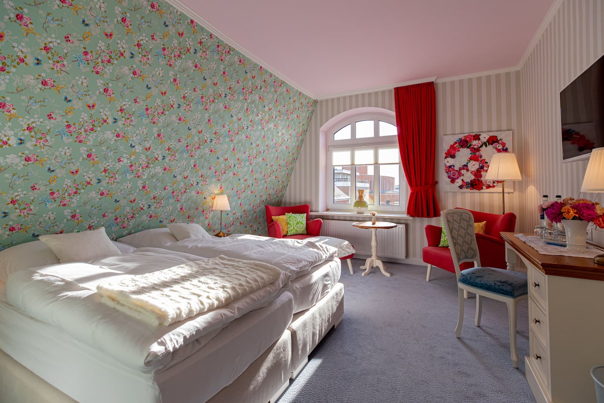 Een slaapkamer met bloemenbehang, een bed met witte lakens, een rode stoel, een bureau met een bloevas en een raam met een rood gordijn.
