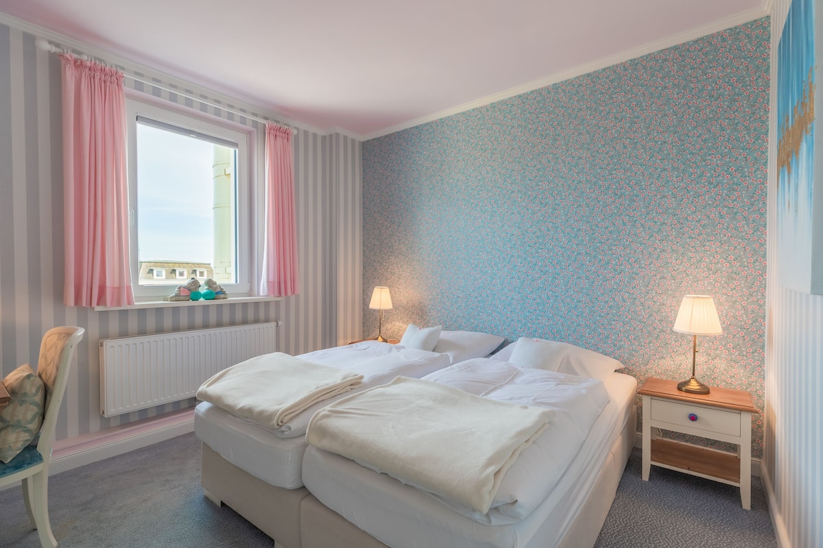 Een slaapkamer met twee bedden, een raam met roze gordijnen, een radiator, een nachtkastje met een lamp en een stoel.