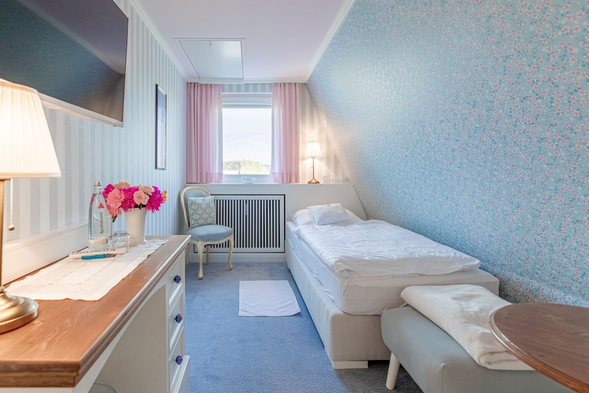 Een hotelkamer met een bed, een stoel, een bureau met lamp, een vaas met bloemen en een raam met gordijnen.