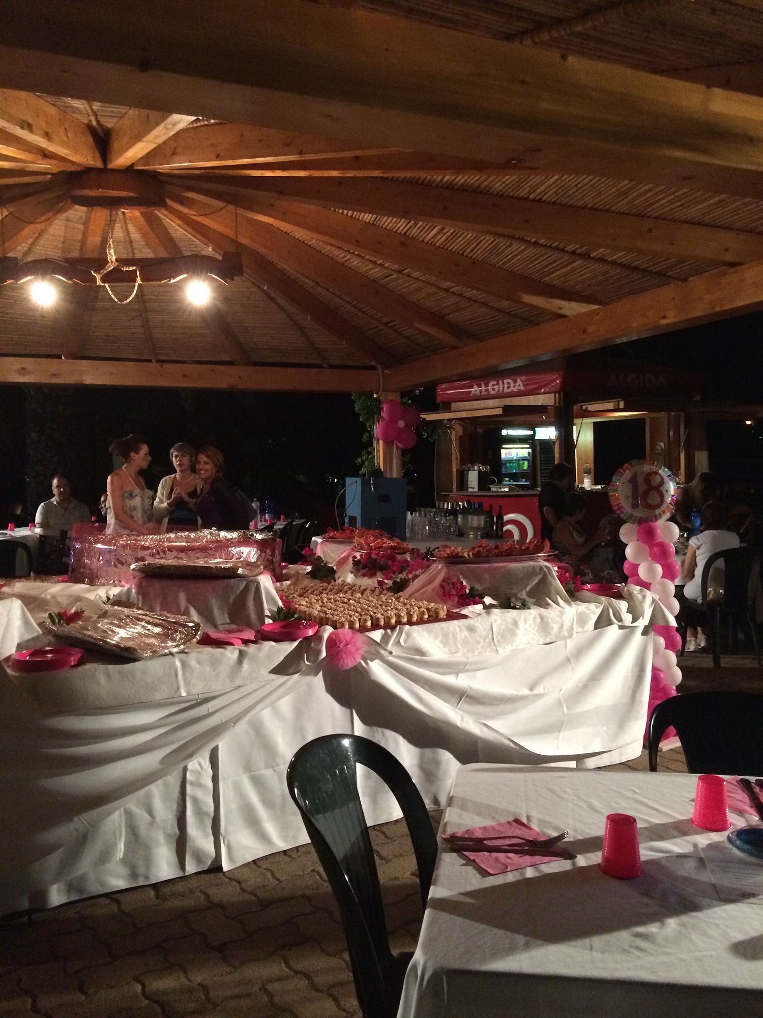 An outdoor dining area with tables covered in white tablecloths, food items, and a bar area in the background.