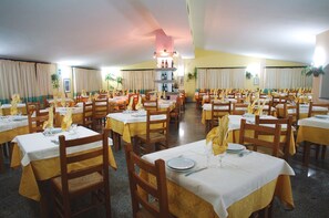 A large dining hall with multiple tables set for service, featuring wooden chairs and white tablecloths. - Le Quattro Lune (Orosei)
