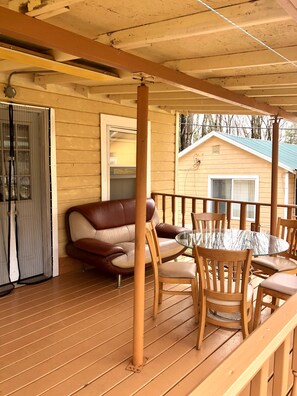 Eine Holzterrasse mit einem Glastisch und Stühlen, ein braunes Ledersofa und ein mit einem Insektengitter versehenes Fenster.